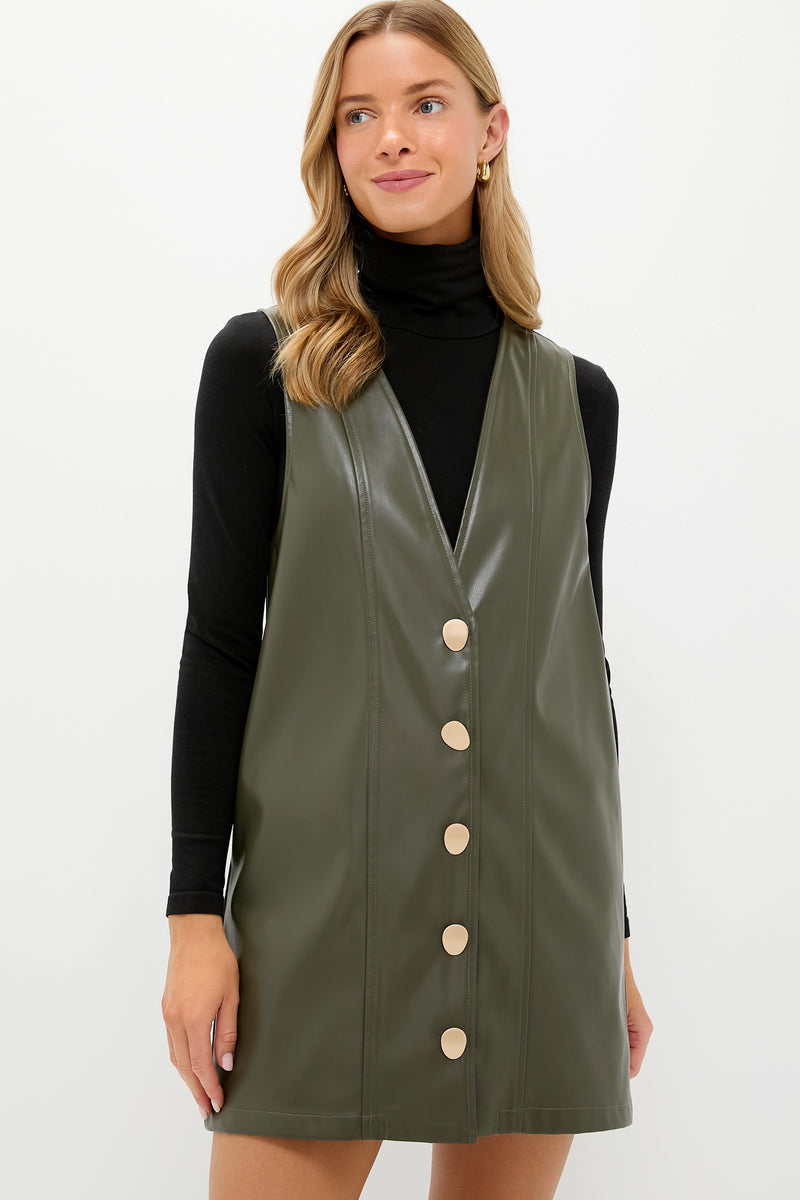 A woman models Hyacinth House’s olive green faux leather pinafore dress with large buttons, styled over a black turtleneck top.