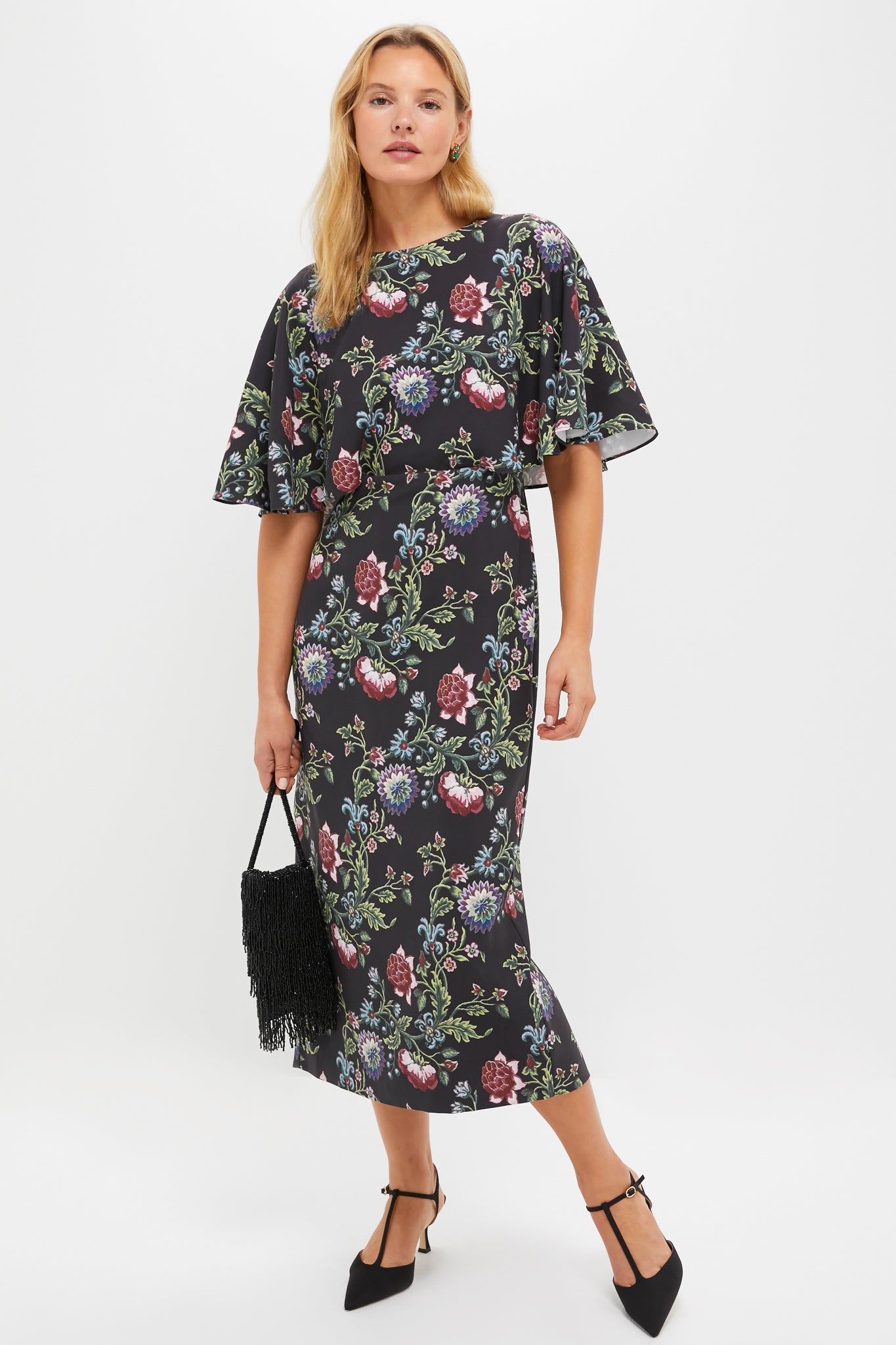 Woman in a classic floral dress, holding a fringed handbag, exemplifying effortless elegance. Pair with Emerald Myrla Earrings for a fun, high-end look.