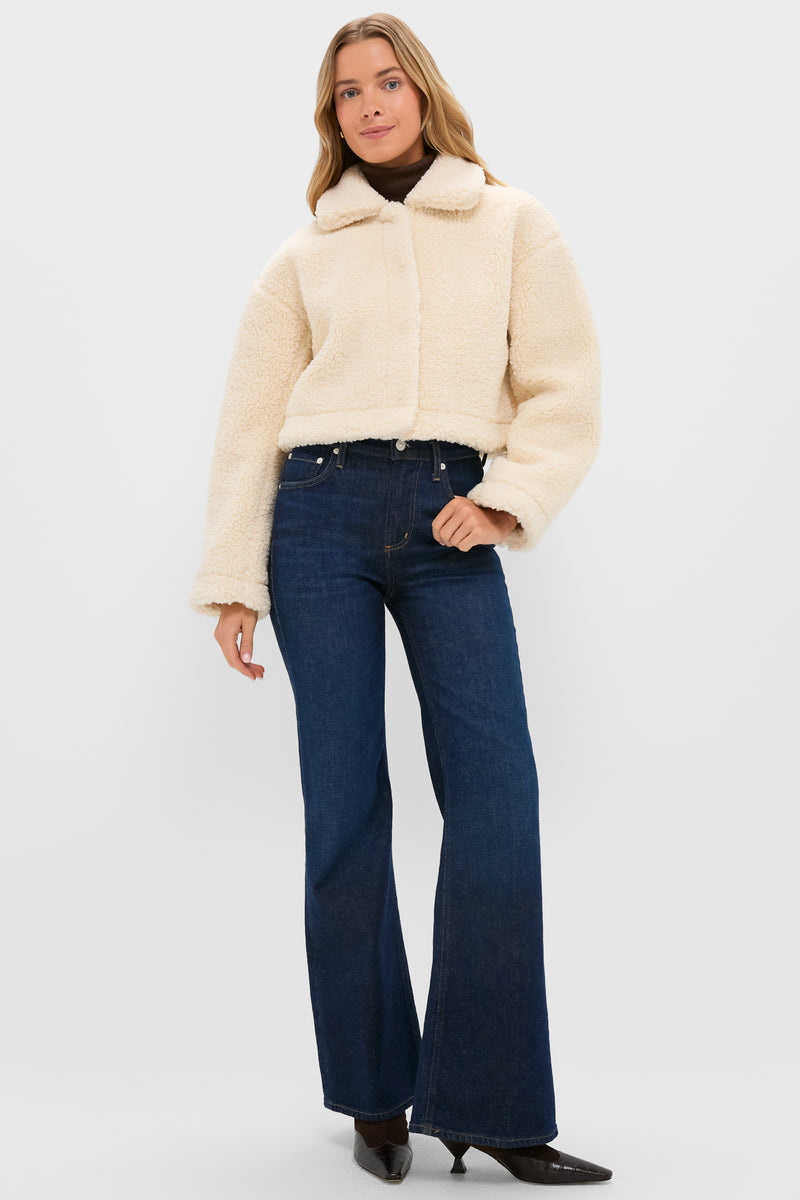 A person in a cream-colored cropped sherpa Sara Jacket by Hyacinth House, dark turtleneck, dark blue flare jeans, and black shoes stands against a light background—an ideal cold-weather style.