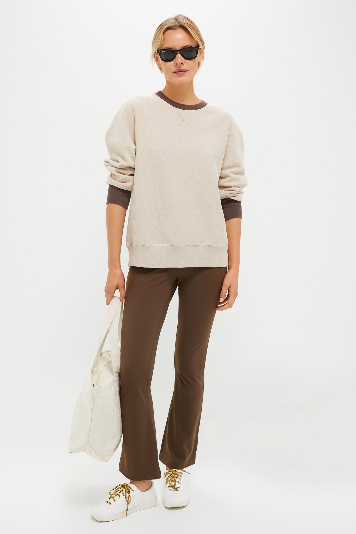 A person in Tnuck Sport sunglasses, a vintage-style crewneck sweatshirt, brown flared pants, and white sneakers holds a white tote bag against a plain background.