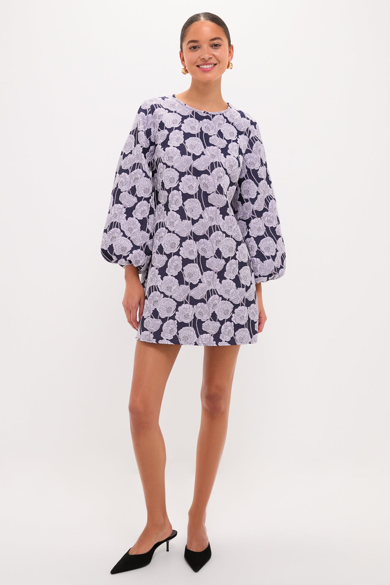 Woman models the classic Lilac & Navy Floral Jacquard Janie Mini Dress, showcasing its effortless elegance with long sleeves, paired with black mules and gold hoops for a fun, high-end look.