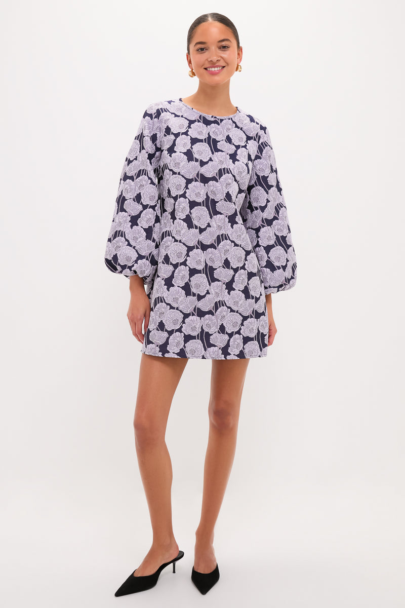 Woman models the classic Lilac & Navy Floral Jacquard Janie Mini Dress, showcasing its effortless elegance with long sleeves, paired with black mules and gold hoops for a fun, high-end look.