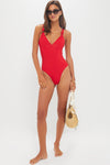 A woman models the stylish Maygel Coronel Vichada One Piece swimsuit with sunglasses and a straw bag, standing barefoot against a plain white backdrop.