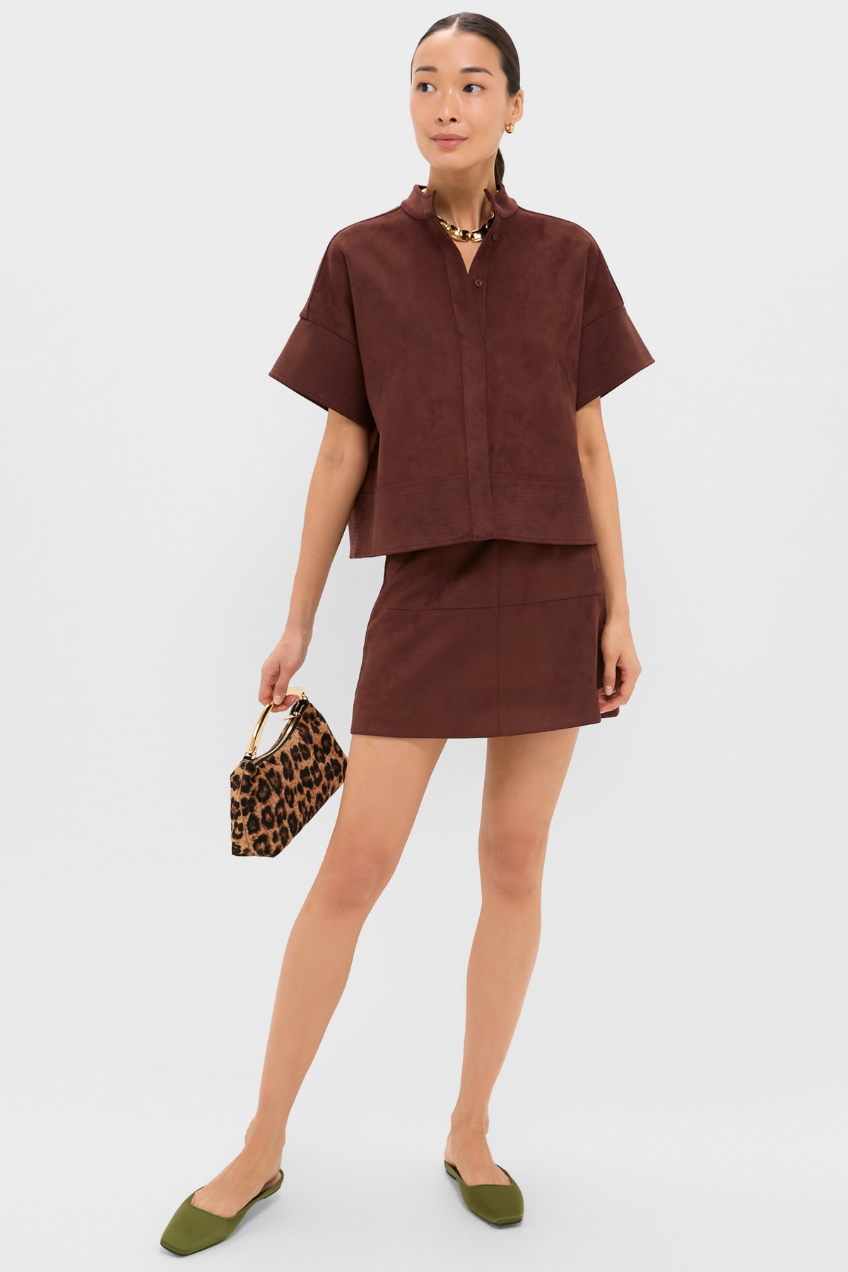 A woman stands against a white background wearing a Tuckernuck Ganache Vegan Suede Shirt with short sleeves and trapunto stitching, paired with a skirt, green flats, and holding a leopard print handbag.
