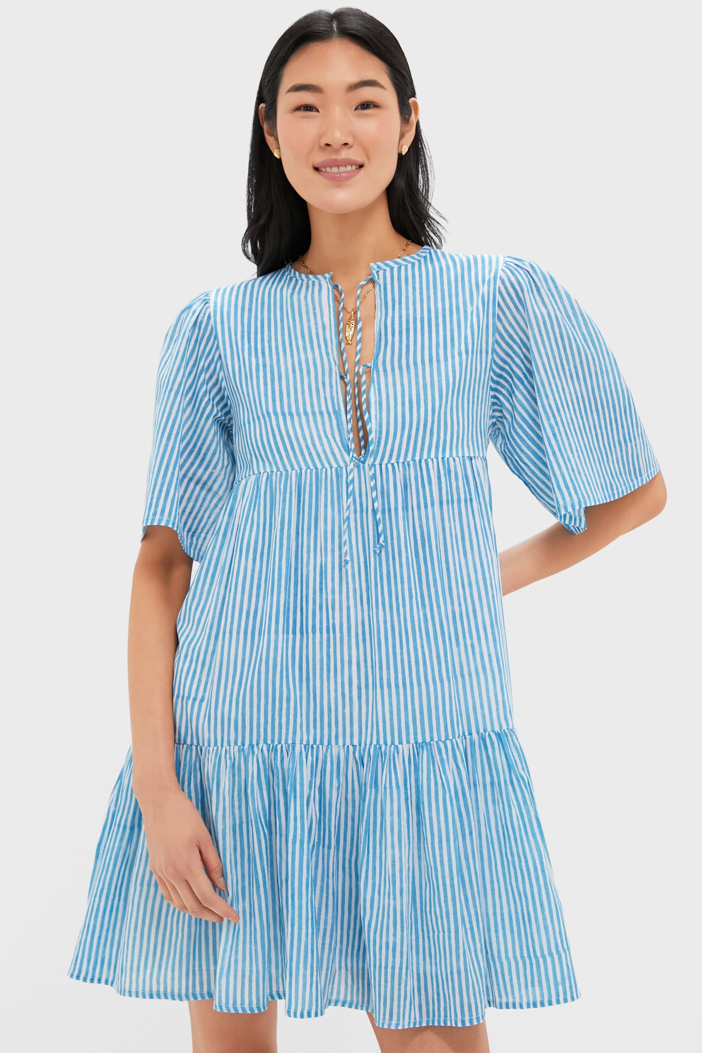 A person stands facing the camera, smiling, in a blue and white vertical striped organic cotton dress with a ruffled hem and lace-up neckline by Love Tomas by Emerson Fry.
