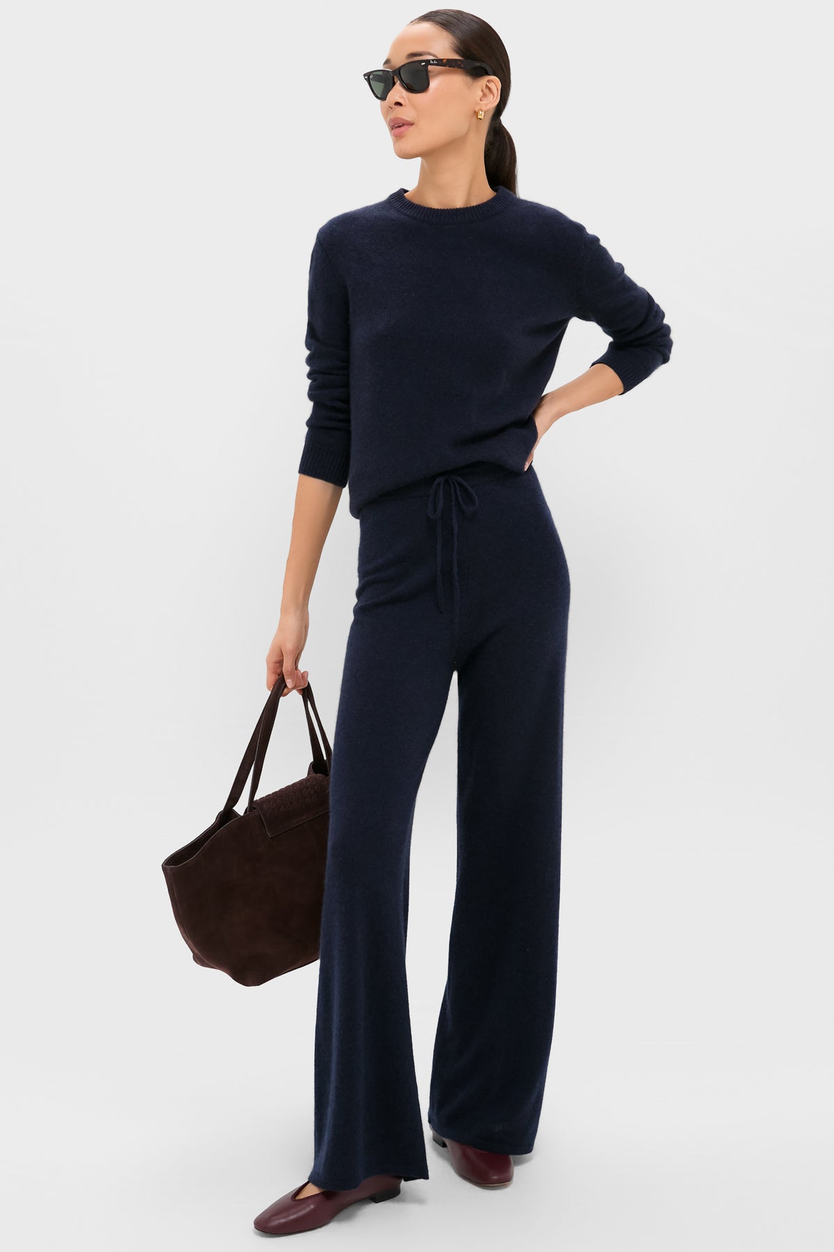 A woman stands against a plain background wearing a Tuckernuck navy blue knit sweater, relaxed fit cashmere pants, burgundy shoes, sunglasses, and holding a dark brown tote bag.
