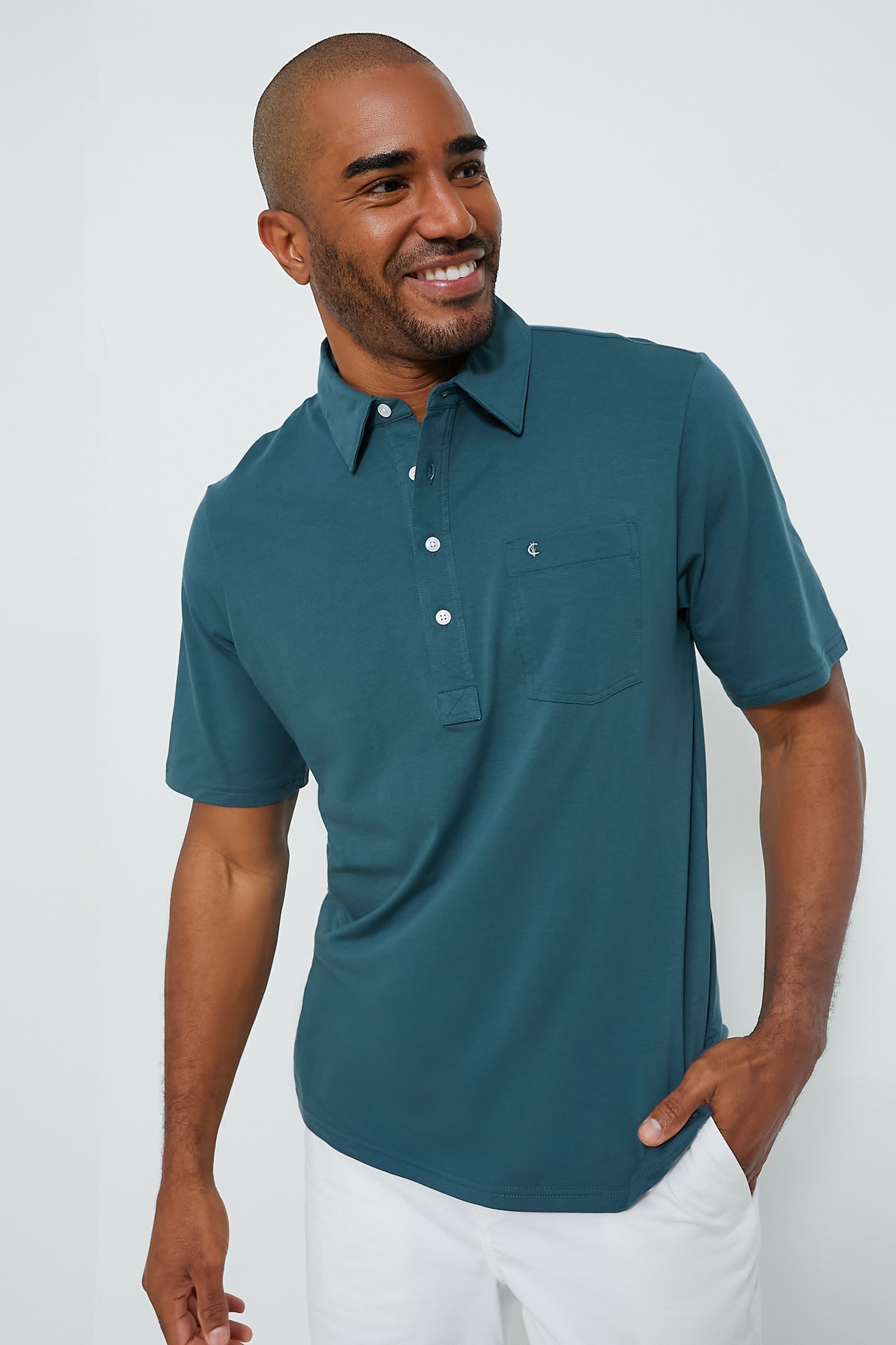 A man smiles with one hand in his pocket, wearing a teal shirt from Criquet and white pants.