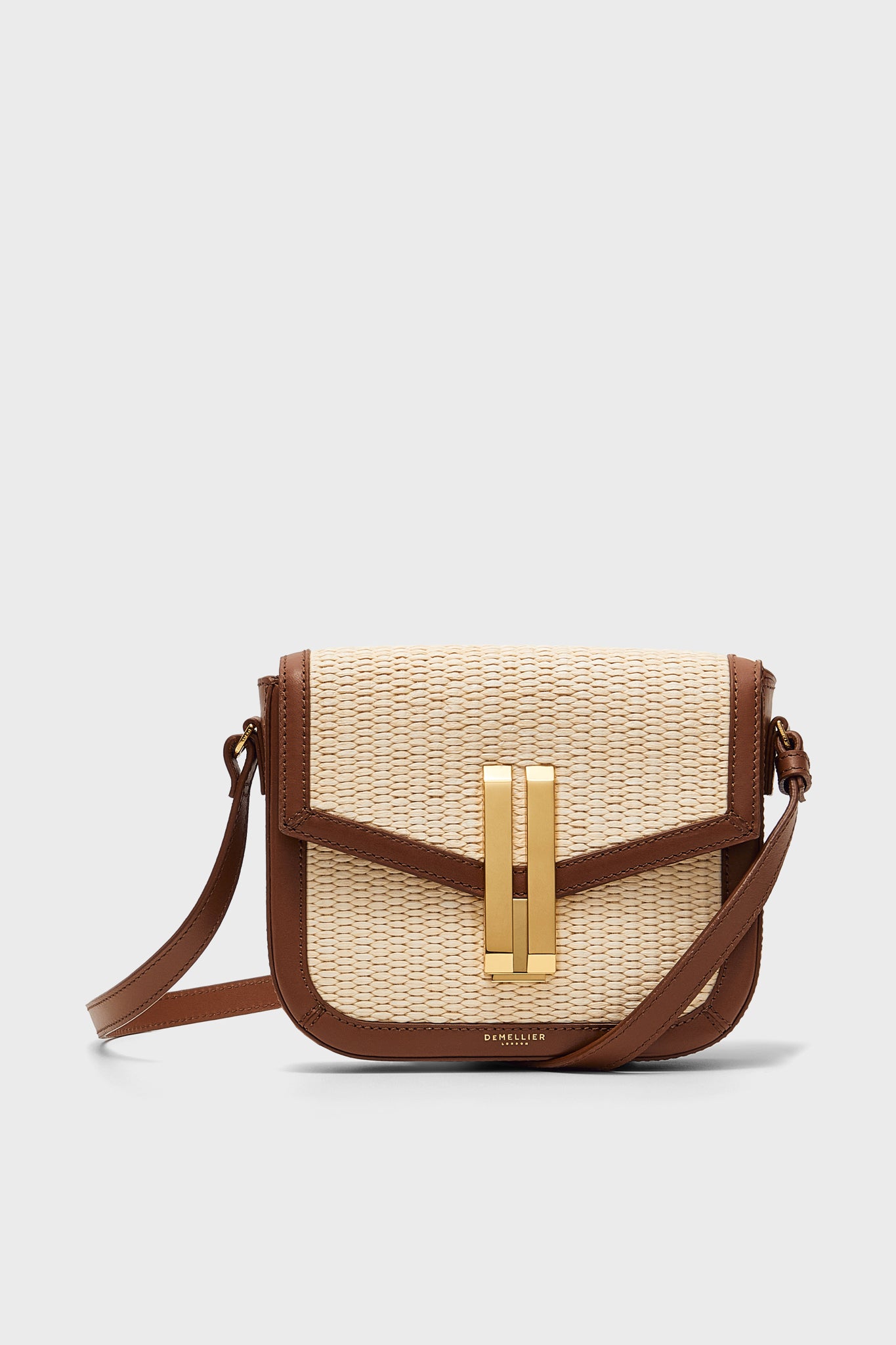 A small Demellier Vancouver crossbody bag with a woven beige front, brown leather trim, and a gold rectangular clasp.