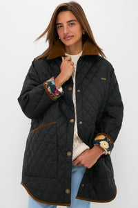 A woman stands against a plain background wearing the Barbour x Tuckernuck Miley quilt jacket with diamond stitching, brown trim, and a corduroy collar over a cream sweater and blue jeans.