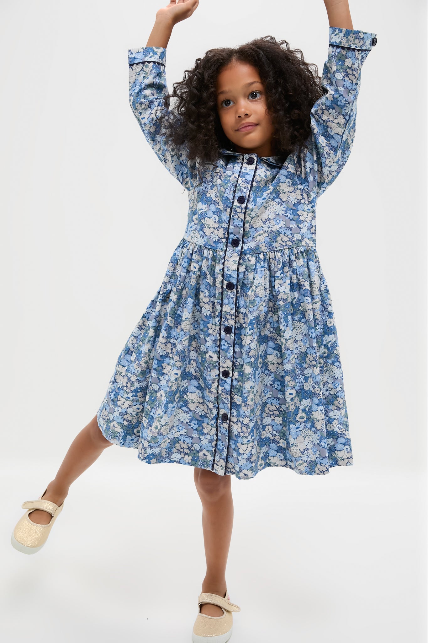Young girl in a classic, long sleeve floral button-front dress, striking a playful pose. Effortless style with frilled collar and ric rac details, paired with fun, glittery shoes.