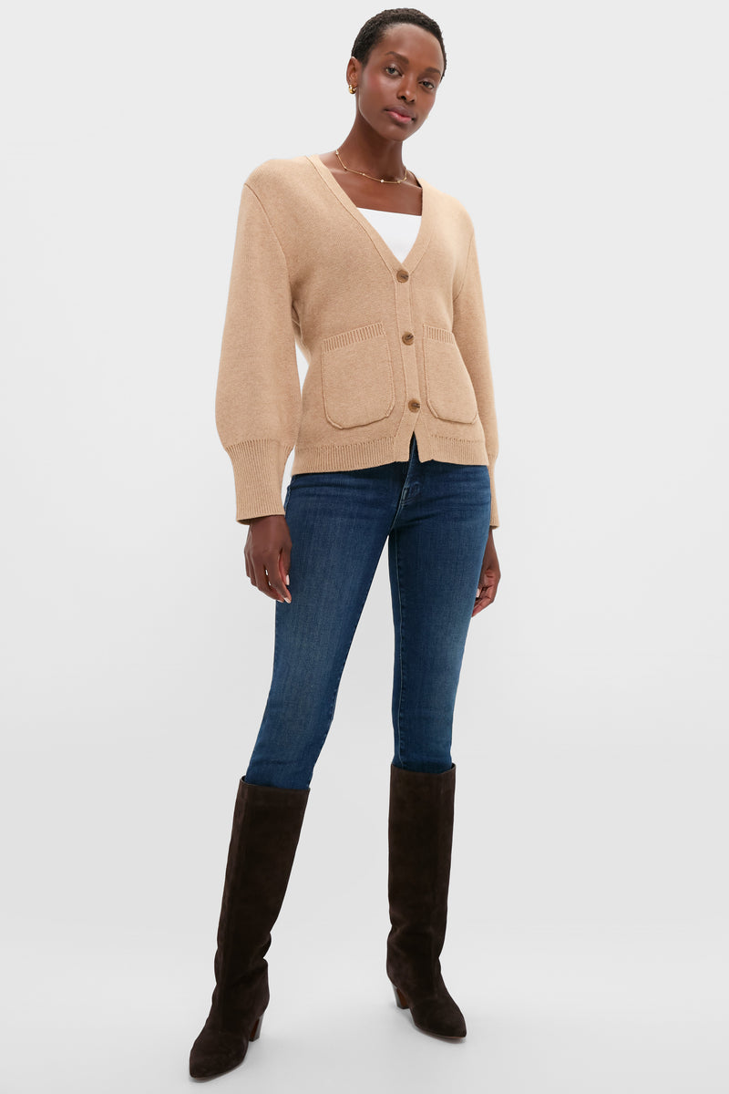 A person stands against a plain white background wearing an ALEXIS beige buttoned cardigan with balloon sleeves, blue jeans, and knee-high black boots.