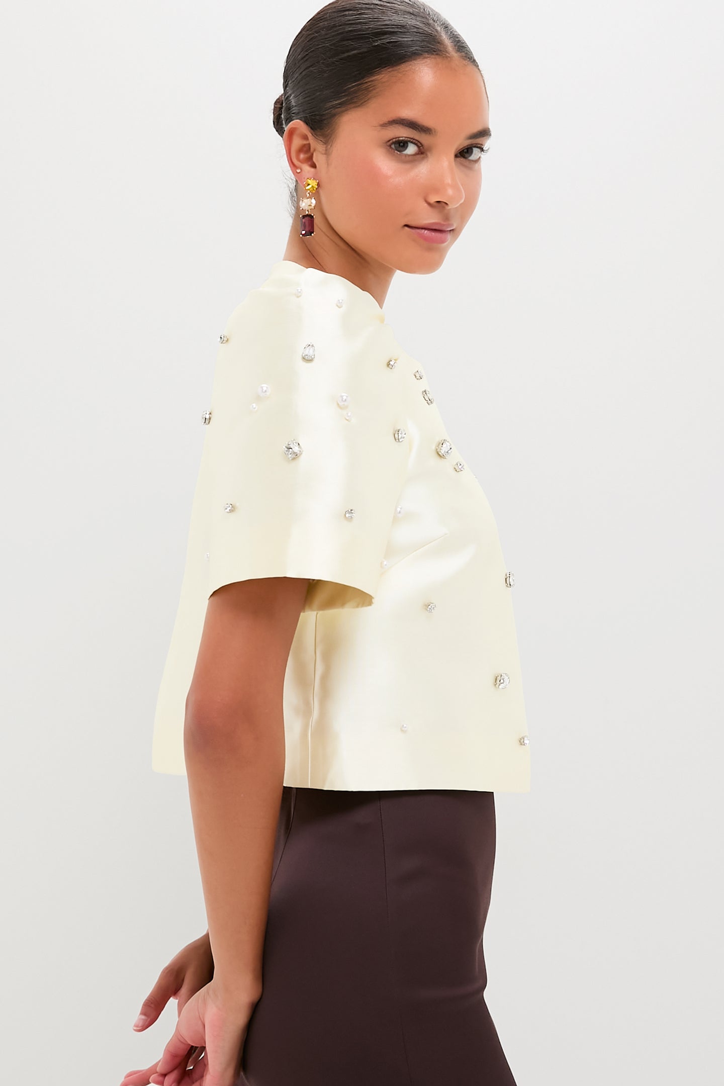 Model in profile wearing the classic Ivory Olivia Top, an effortless high-end cropped satin design with fun pearl and rhinestone embellishments, paired with a fitted dark-brown skirt.