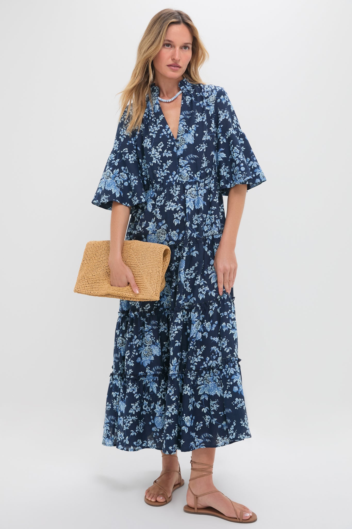 A woman in Tuckernucks Twilight Garden Jenna Dress—a navy blue floral midi perfect for spring—holds a woven clutch and stands in tan flat sandals against a plain white background.