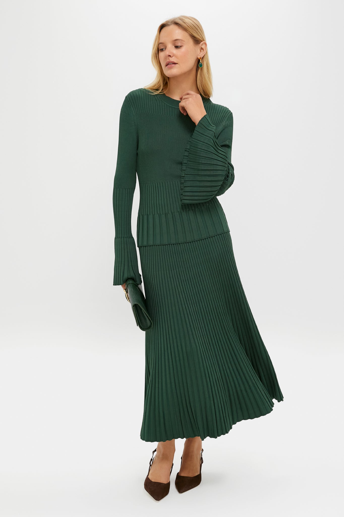 A woman wears a Zimmermann dark green ribbed knit sweater dress with bell sleeves and a pleated skirt, styled with a matching green clutch and brown heels.