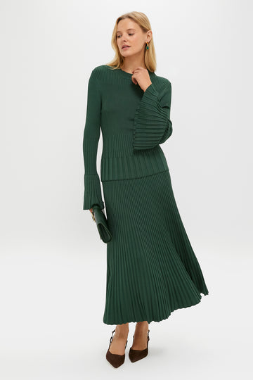 A woman wears a Zimmermann dark green ribbed knit sweater dress with bell sleeves and a pleated skirt, styled with a matching green clutch and brown heels.