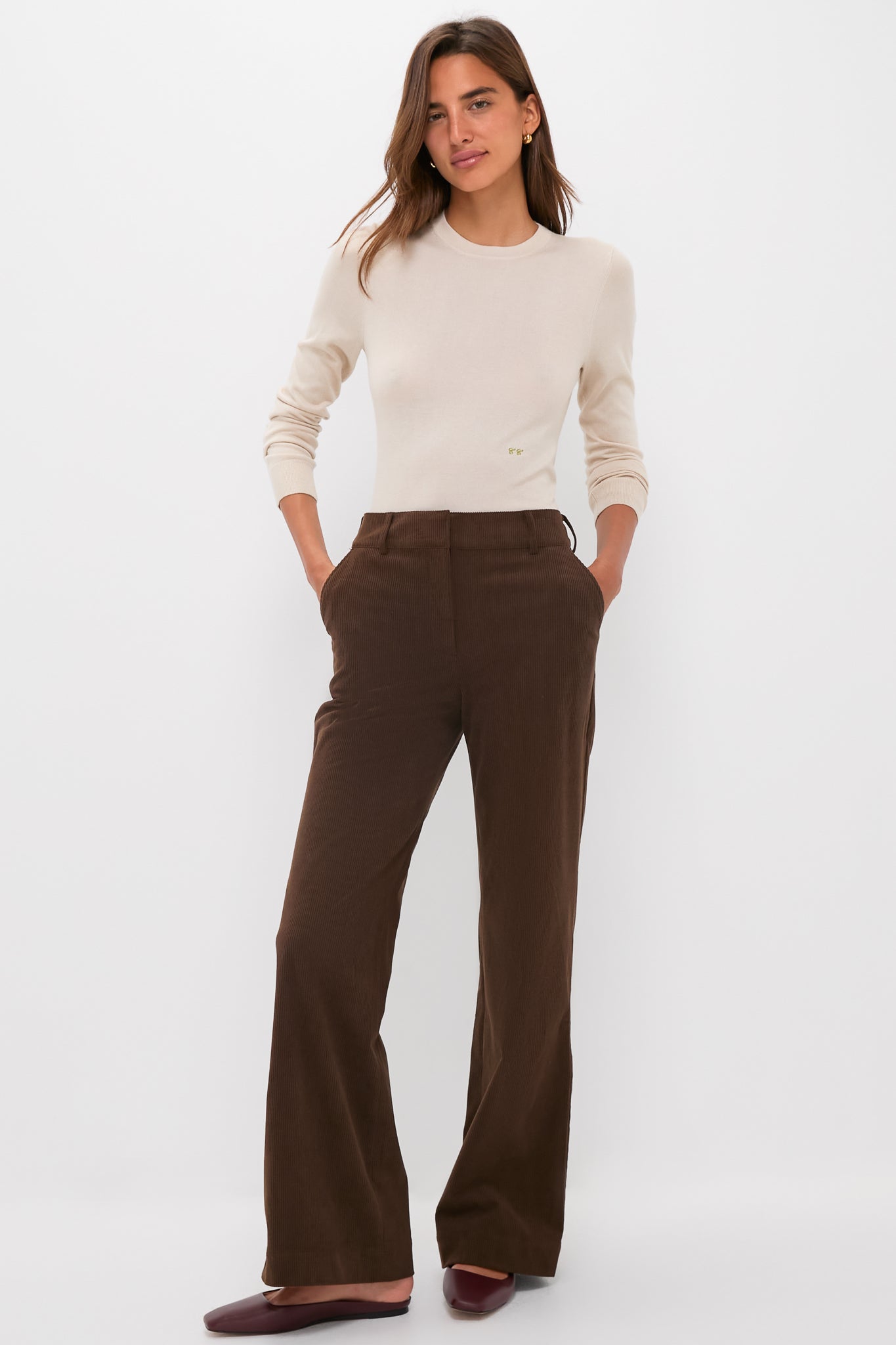 A woman stands against a white background wearing a Tuckernuck cream crewneck cashmere silk tee, high-waisted brown wide-leg pants, and burgundy flats, with her hands in her pockets.