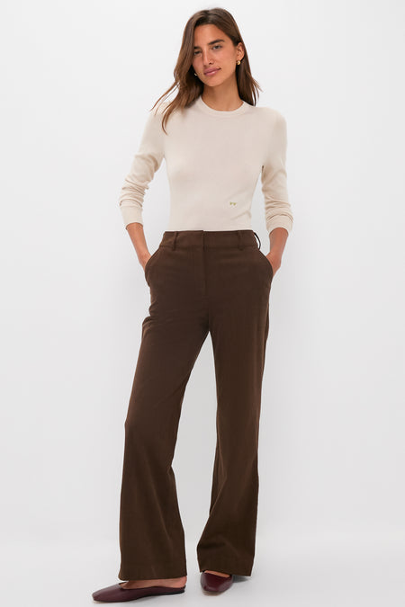 A woman stands against a white background wearing a Tuckernuck cream crewneck cashmere silk tee, high-waisted brown wide-leg pants, and burgundy flats, with her hands in her pockets.