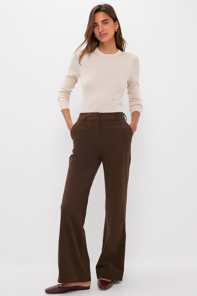 A woman stands against a white background wearing a Tuckernuck cream crewneck cashmere silk tee, high-waisted brown wide-leg pants, and burgundy flats, with her hands in her pockets.