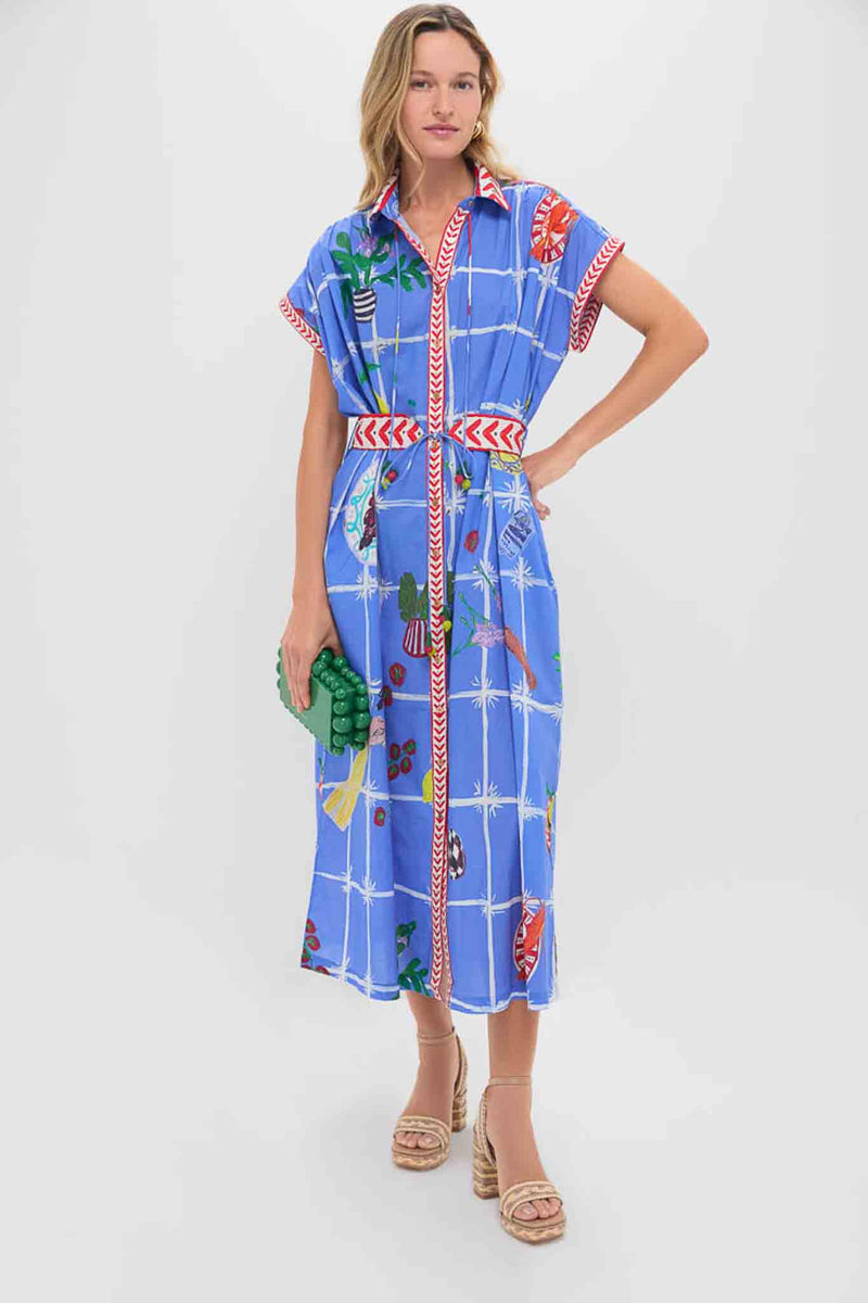 A woman wears a Carolina K. blue belted waist dress with a colorful picnic-inspired pattern and red belt, holding a green clutch and wearing platform sandals.