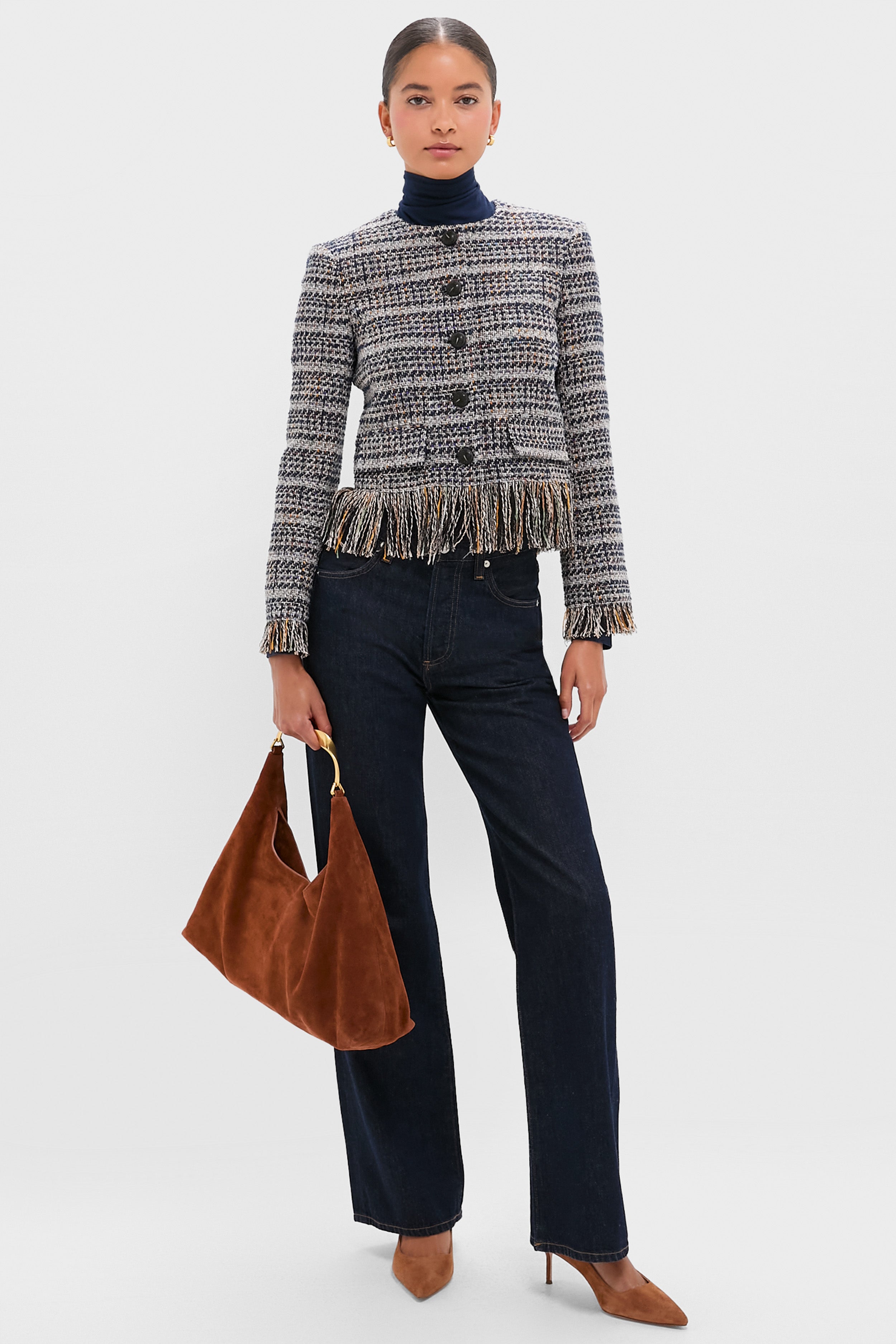 A woman stands wearing a Veronica Beard tweed jacket with fringe, dark wide-leg jeans, brown heels, and holding a brown suede handbag.