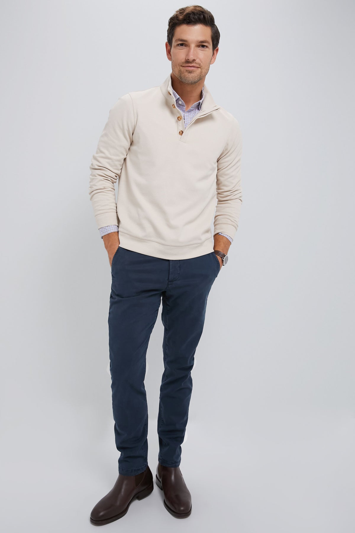 A man stands against a plain background wearing Spruce’s light beige cotton sweater, patterned shirt, navy pants, and brown boots—hands in his pockets and looking at the camera—highlighting comfortable essentials for a versatile wardrobe.