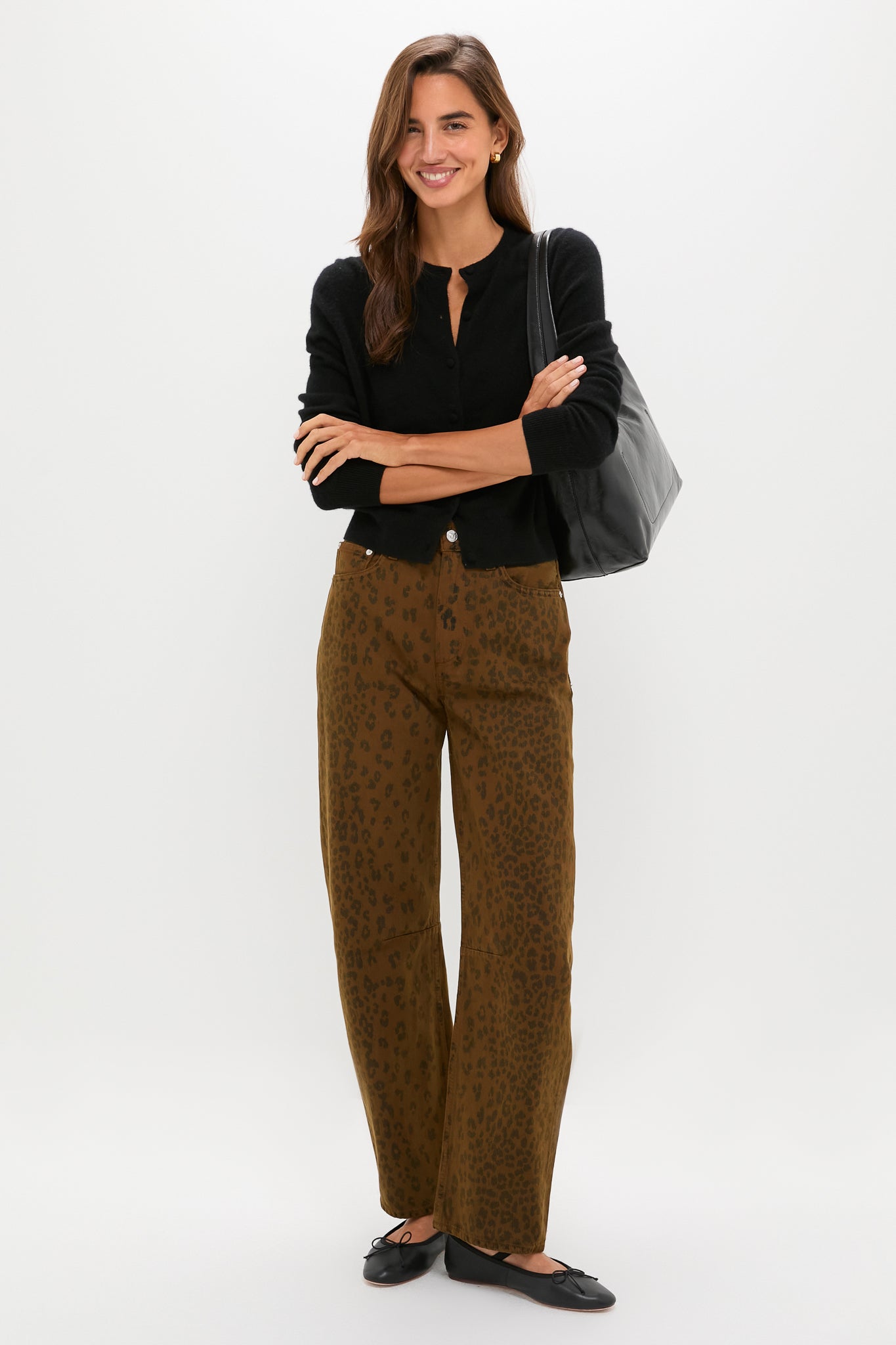 A woman stands against a white background wearing cheetah print pants by Citizens of Humanity, a black cardigan, black flats, and a black shoulder bag—her bold style offers an elevated everyday look with chic jean alternatives.