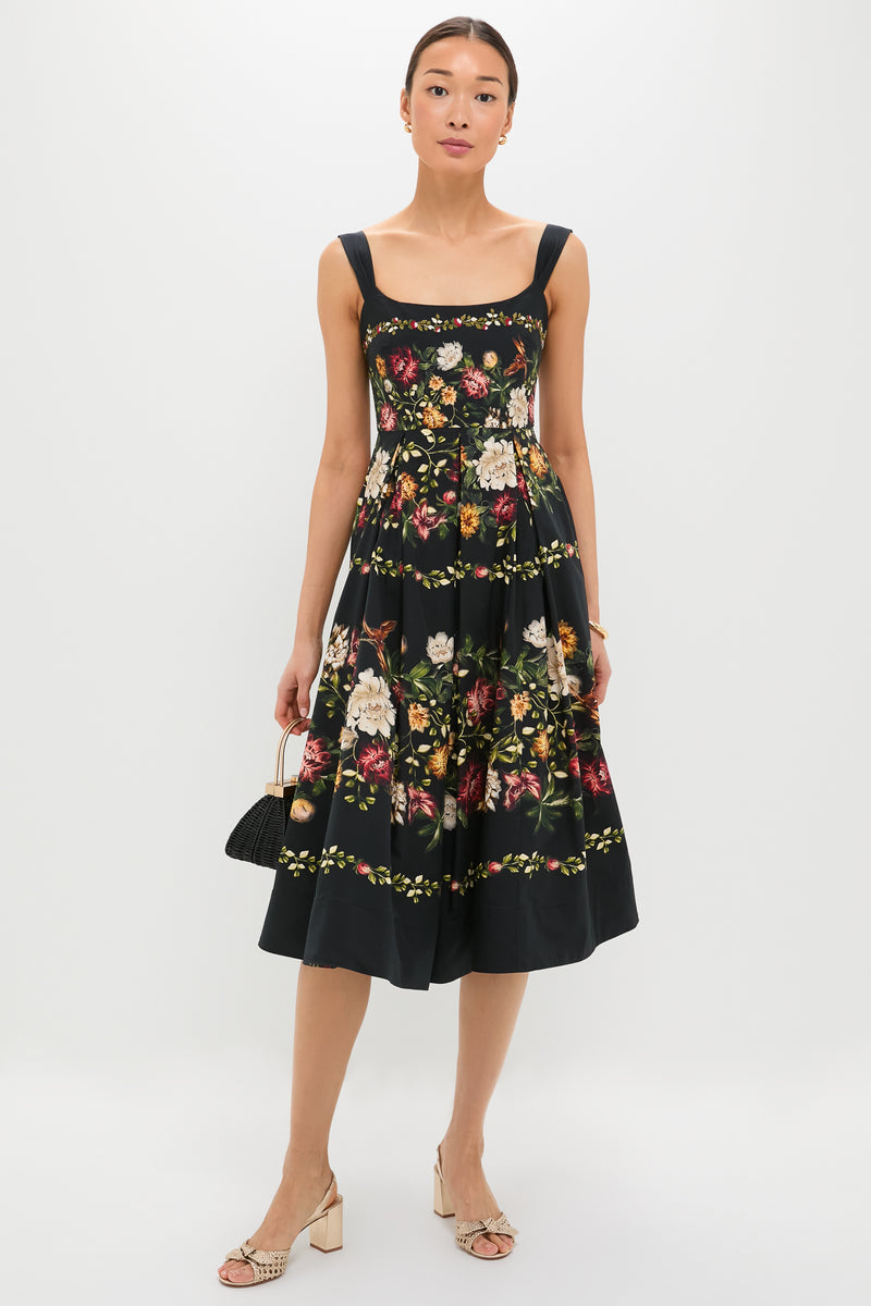 Model wears the Paramo Alcaparra Midi Dress, a classic sleeveless black dress with vibrant floral print, paired with a fun, structured handbag and effortless woven-strap sandals.