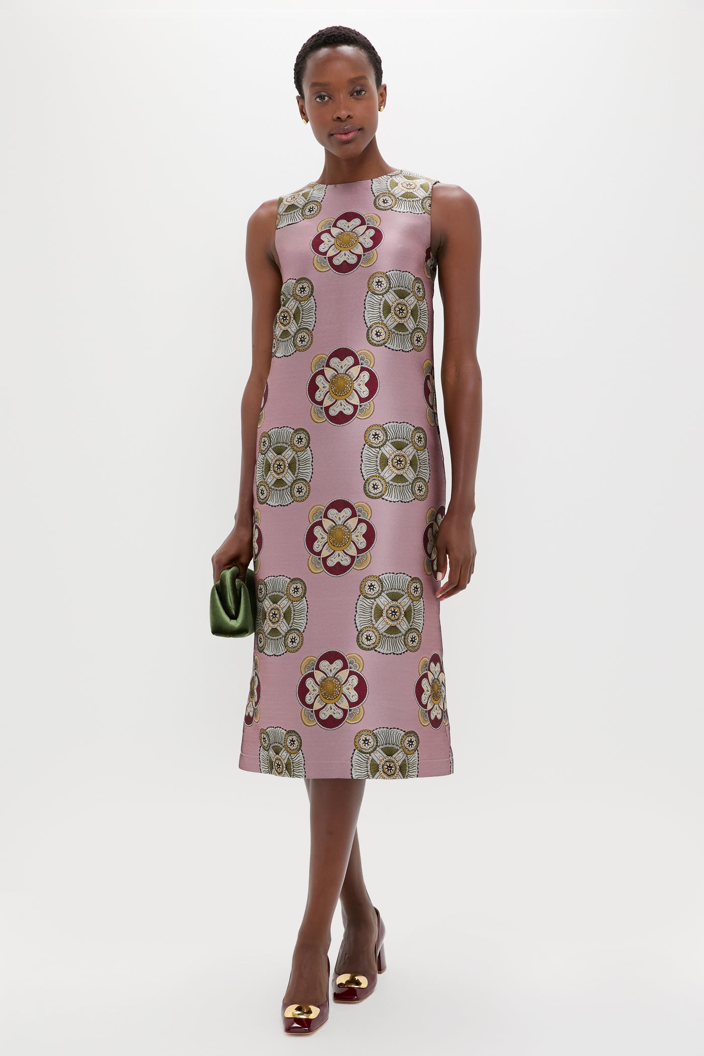 A woman in a sleeveless pink jacquard floral dress by La DoubleJ holds a small green clutch, standing against a plain white background.
