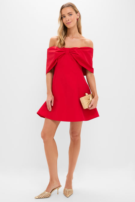 A woman in a Hyacinth House short, off-the-shoulder red dress with a large bow—perfect as a mini holiday look—holds a beige clutch and wears gold striped heels, standing against a plain white background.