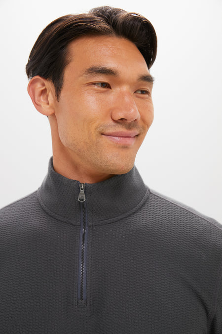 A man with short dark hair smiles slightly, wearing a Faherty charcoal gray textured cotton quarter-zip pullover sweater with a high collar, against a plain white background.
