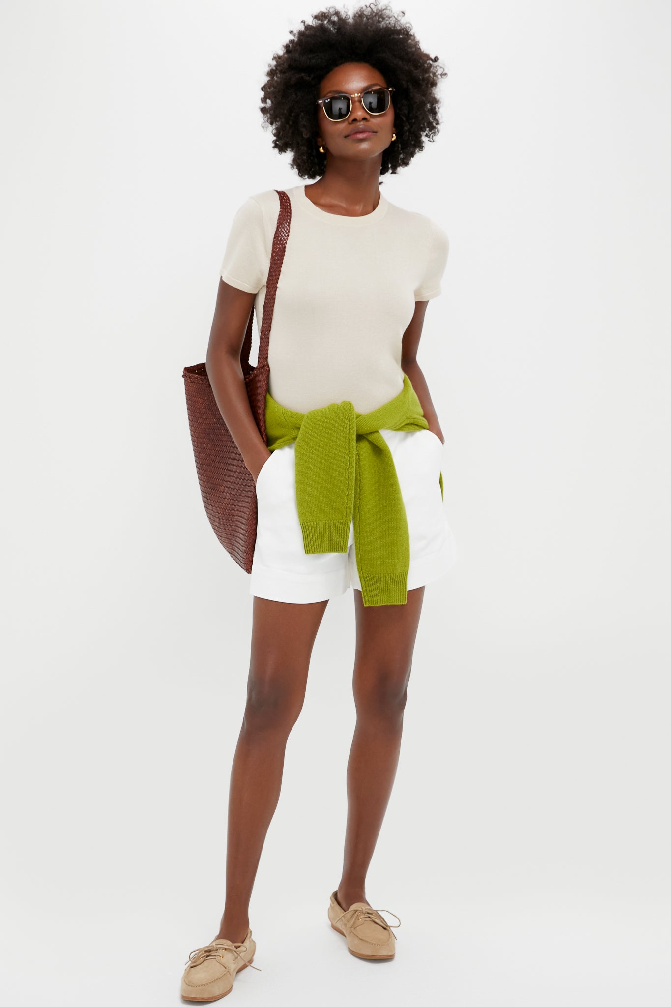 A person in Tuckernuck Ruth Shorts, sunglasses, a beige t-shirt, tan shoes, and a green sweater tied at the waist stands against a white background holding a brown woven bag.
