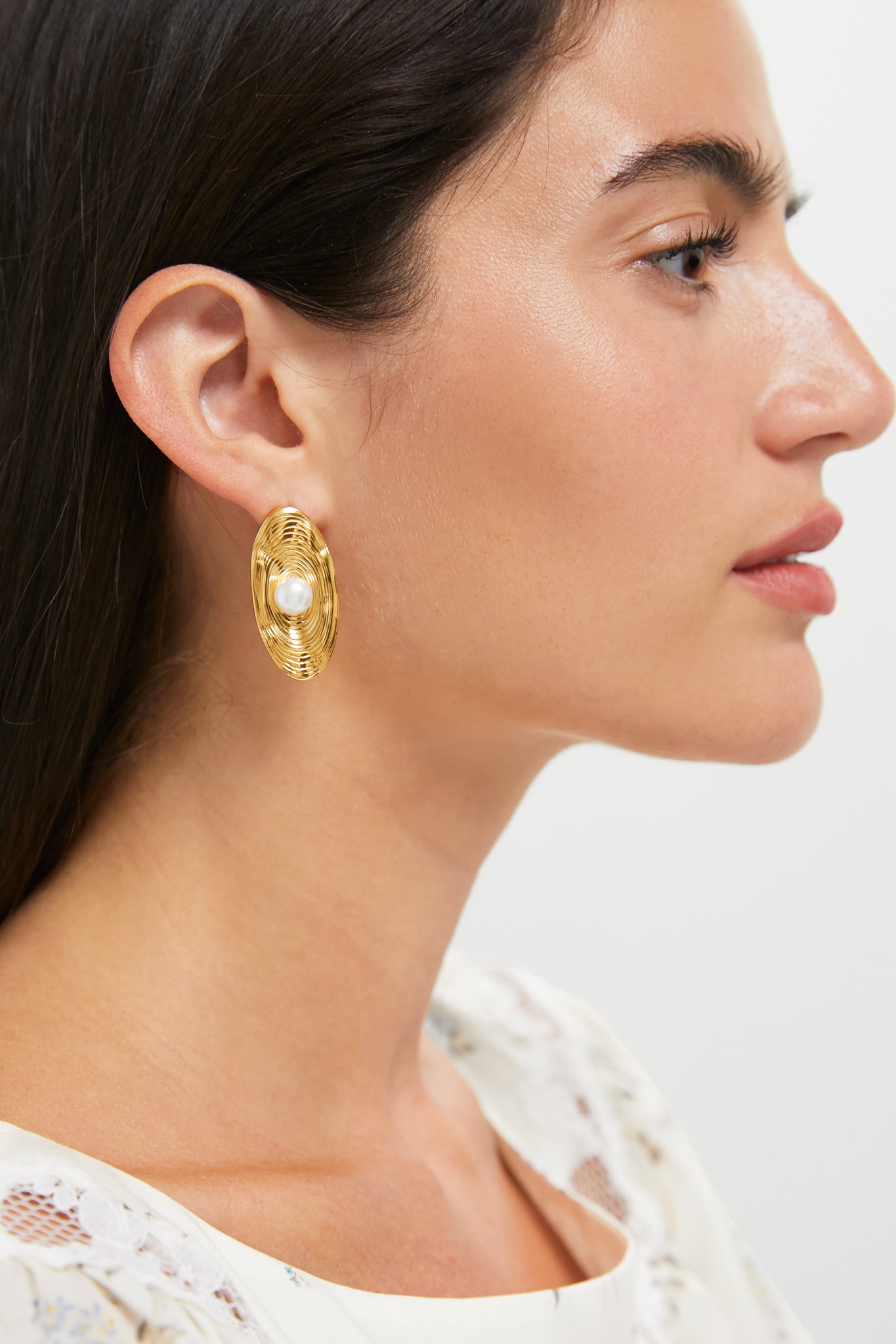 Side profile of a woman wearing Gold Trumbull Earrings: classic gold oval studs with a central pearl, exuding effortless elegance and high-end style.