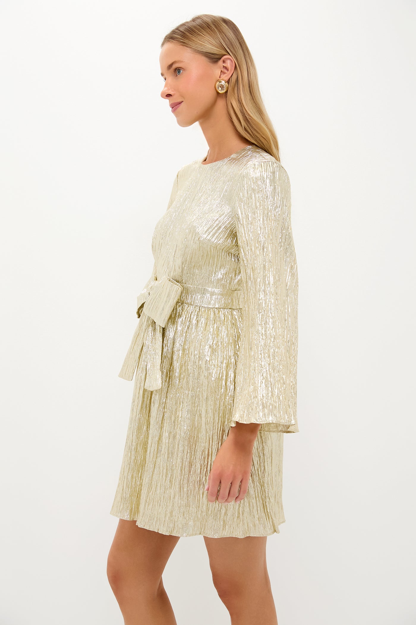Young woman in a studio wearing the Gold Plisse Carol Bow Mini Dress, showcasing its classic, high-end appeal with effortless, fun pleats and a flattering silhouette.