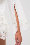 A close-up of a persons arm wearing a Sea New York white blouse featuring lace details, a sleeve slit, and delicate soutache embroidery.