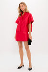 A woman models a Tuckernuck bright red Dupioni top with a boxy silhouette and matching skirt, paired with a black clutch and black pointed-toe flats featuring silver detailing, in a simple studio background.