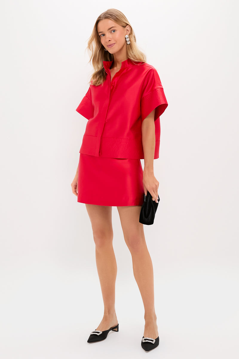 A woman models a Tuckernuck bright red Dupioni top with a boxy silhouette and matching skirt, paired with a black clutch and black pointed-toe flats featuring silver detailing, in a simple studio background.