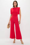 Woman in a sleek, vibrant red jumpsuit, accessorized with an elegant beige clutch and gold-tone mules, embodying a classic, effortless, and high-end style.