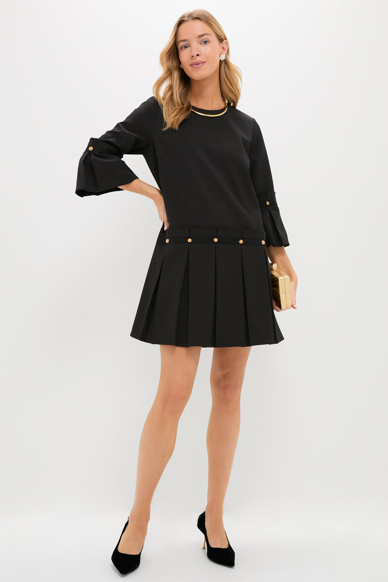 A woman stands against a white background wearing Tuckernuck's Kennedy Dress—a chic little black dress with a pleated skirt, gold button details, black heels, and a gold clutch.