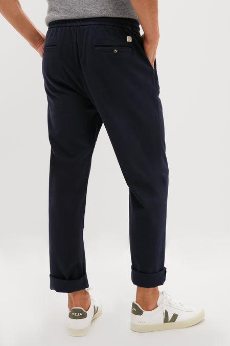 A person wearing Marine Layer’s sustainably-made navy blue Saturday Pant, a grey shirt, and white Veja sneakers stands with hands in pockets, back to camera, against a plain background.