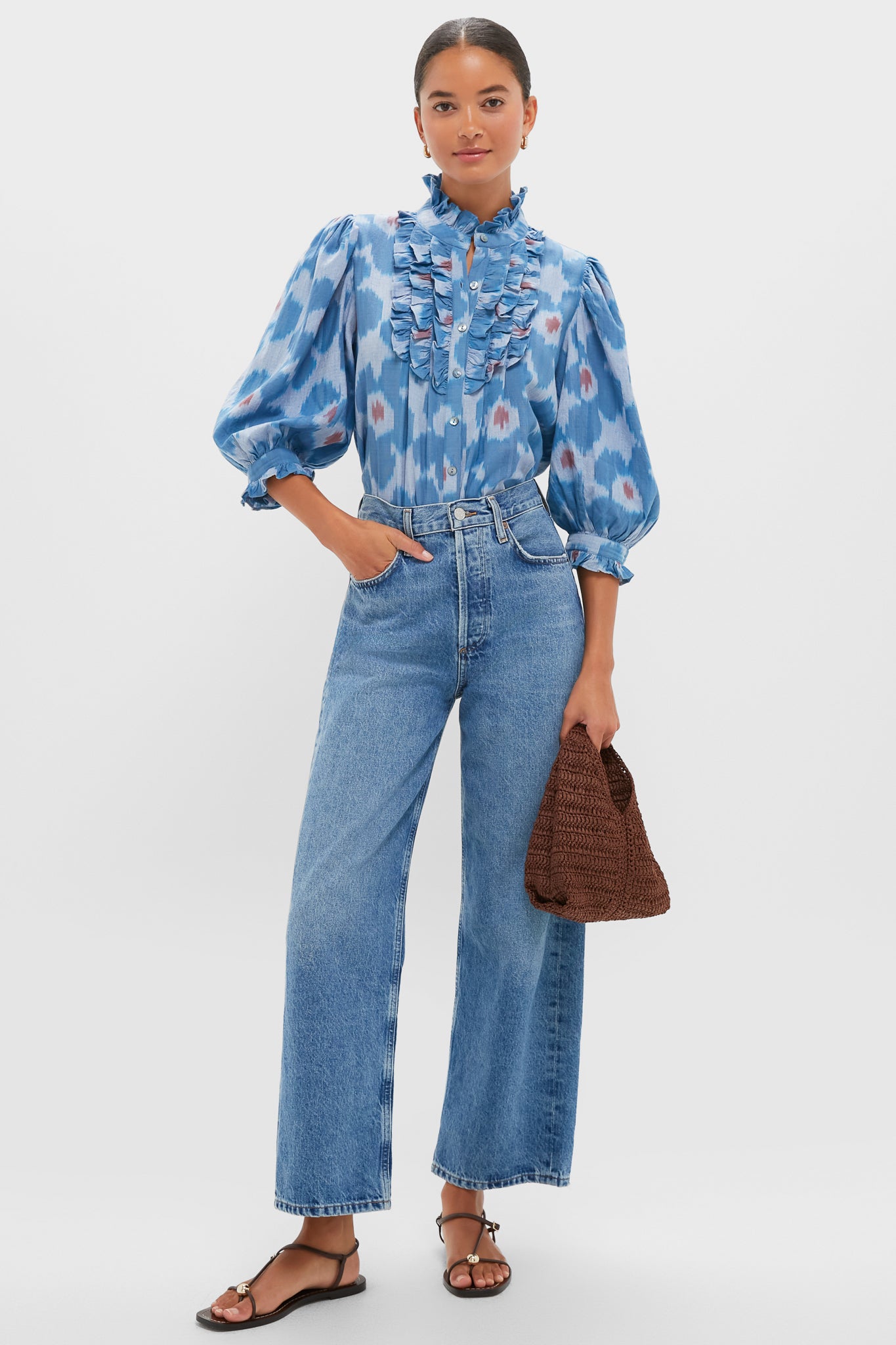 A woman stands wearing the Oliphant Jinja Blue Blouse with ruffle bib detail, high-waisted wide-leg jeans, black sandals, and holding a brown woven bag.