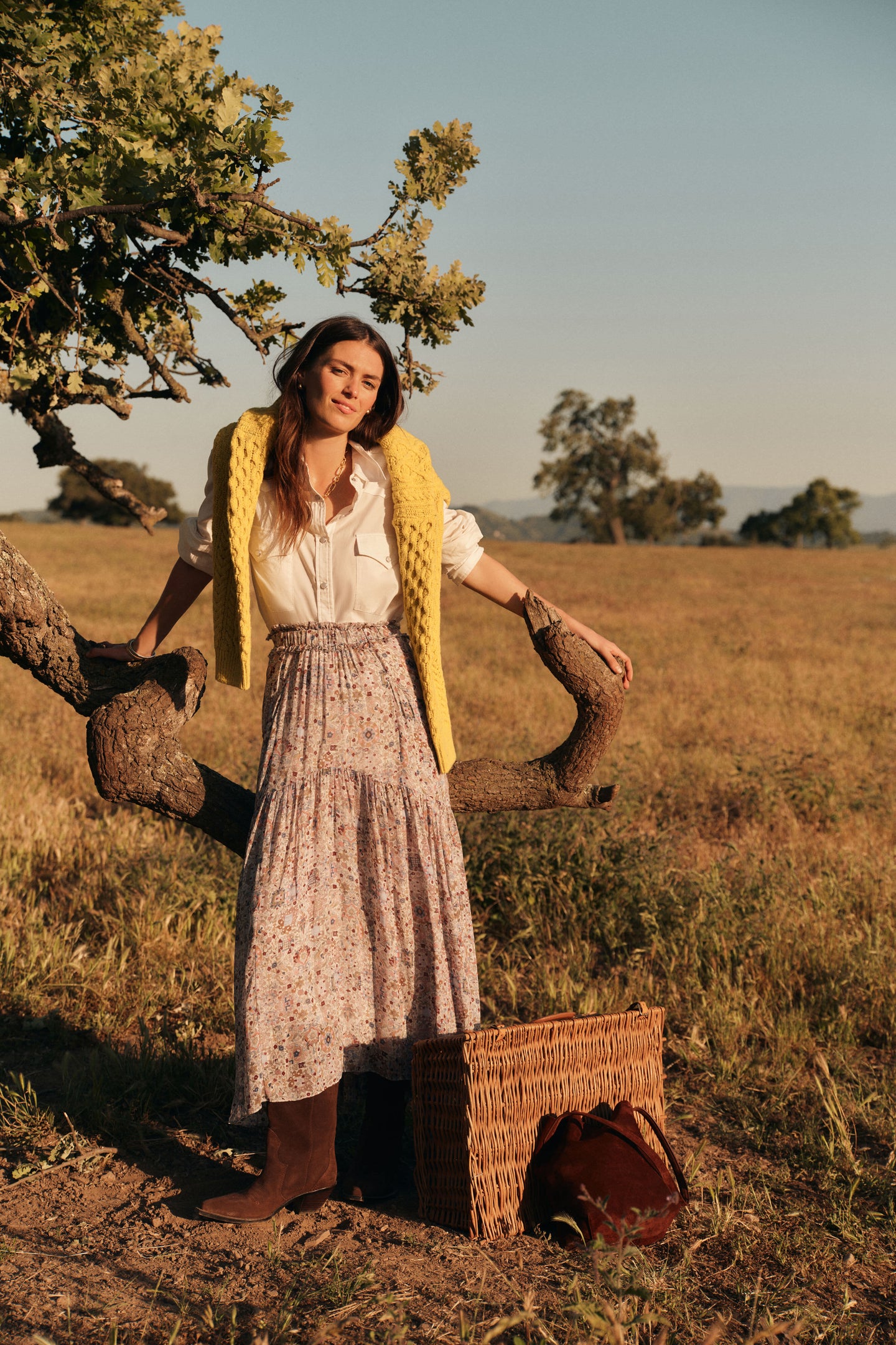 Woman standing outside in a long sleeved white button down shirt with the sleeves rolled up, a yellow cable knit sweater around her shoulders, a floral patterned maxi skirt, and chocolate brown boots. 