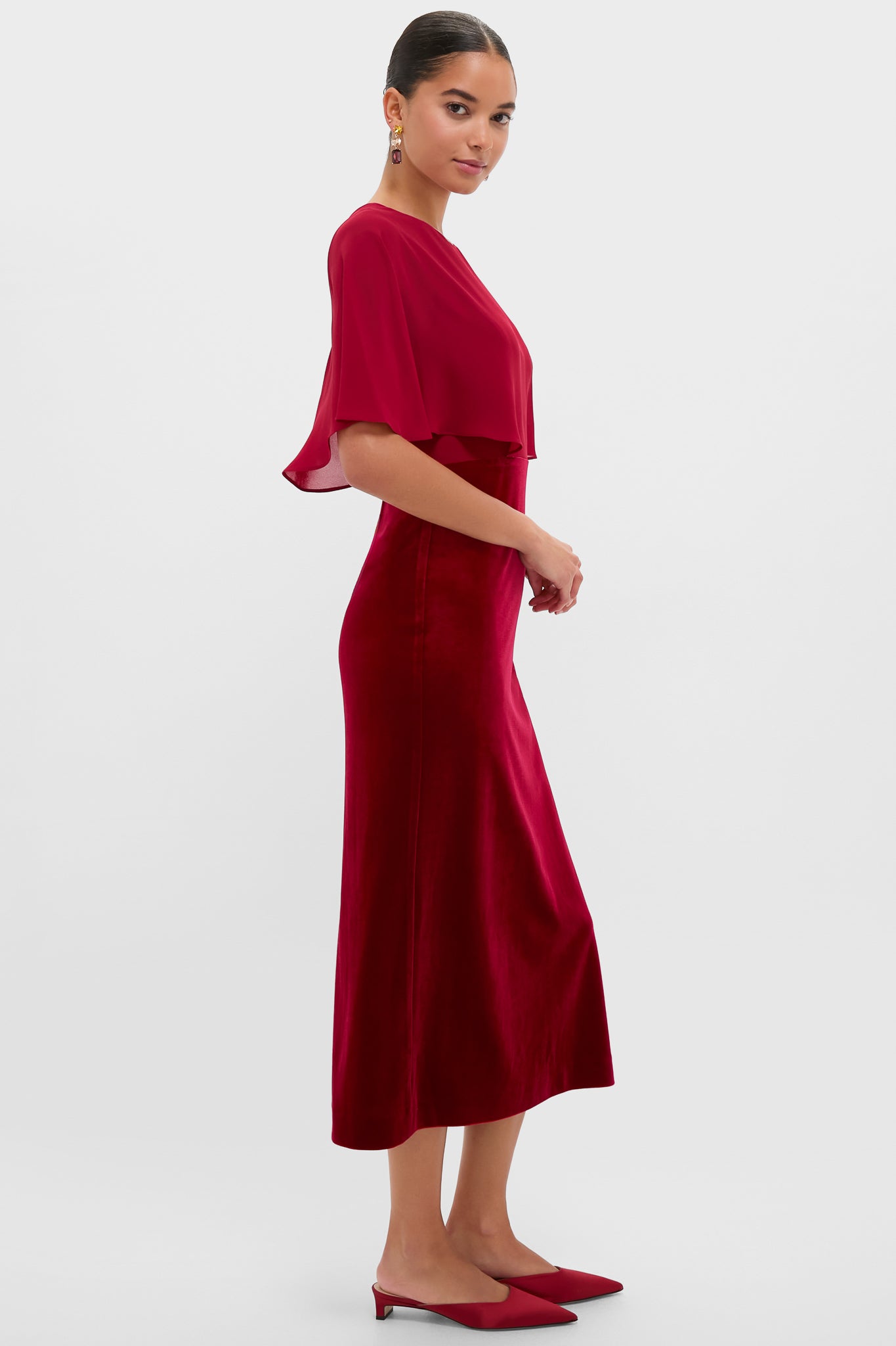Model in profile wearing classic Cranberry Velvet Chiffon Mixed Media Cape Leah Dress, featuring an effortless capelet top, velvet skirt, and matching red mules, exuding high-end, fun elegance.