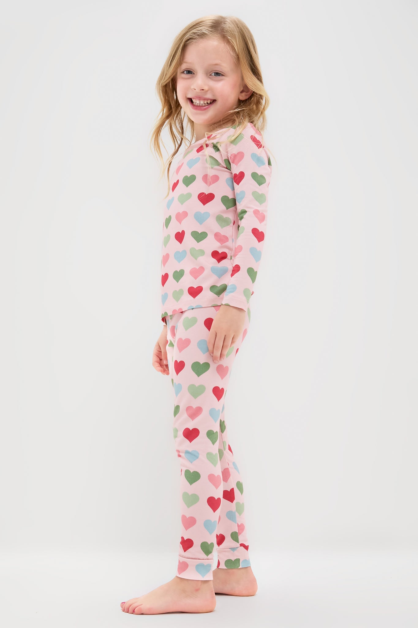 A young child stands barefoot, smiling, wearing Pink Chicken light pink two-piece kids sleepwear pajamas with colorful hearts, against a plain white background.