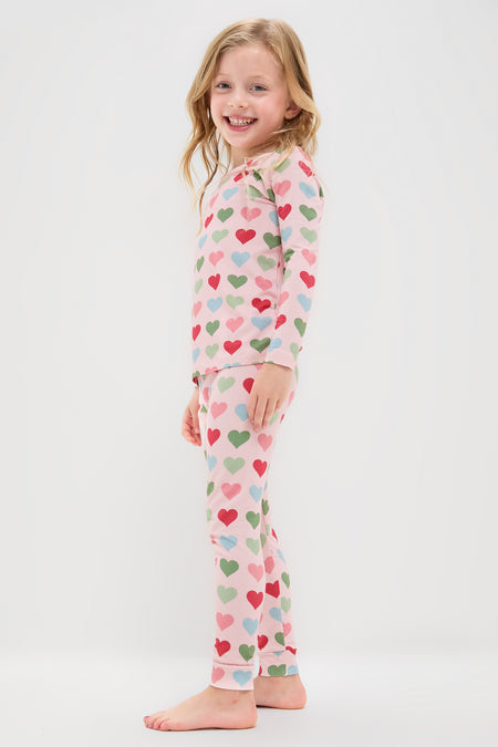 A young child stands barefoot, smiling, wearing Pink Chicken light pink two-piece kids sleepwear pajamas with colorful hearts, against a plain white background.