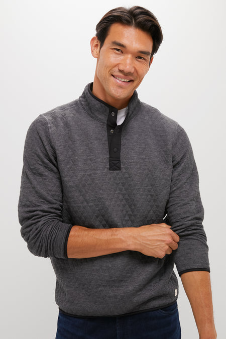 A man with dark hair smiles, wearing a Marine Layer quilted quarter snap gray pullover and dark jeans, one arm crossed over his body.