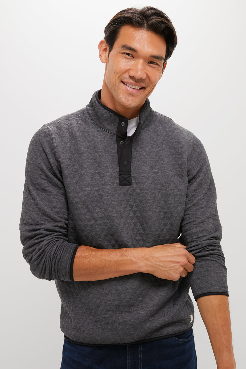 A man with dark hair smiles, wearing a Marine Layer quilted quarter snap gray pullover and dark jeans, one arm crossed over his body.