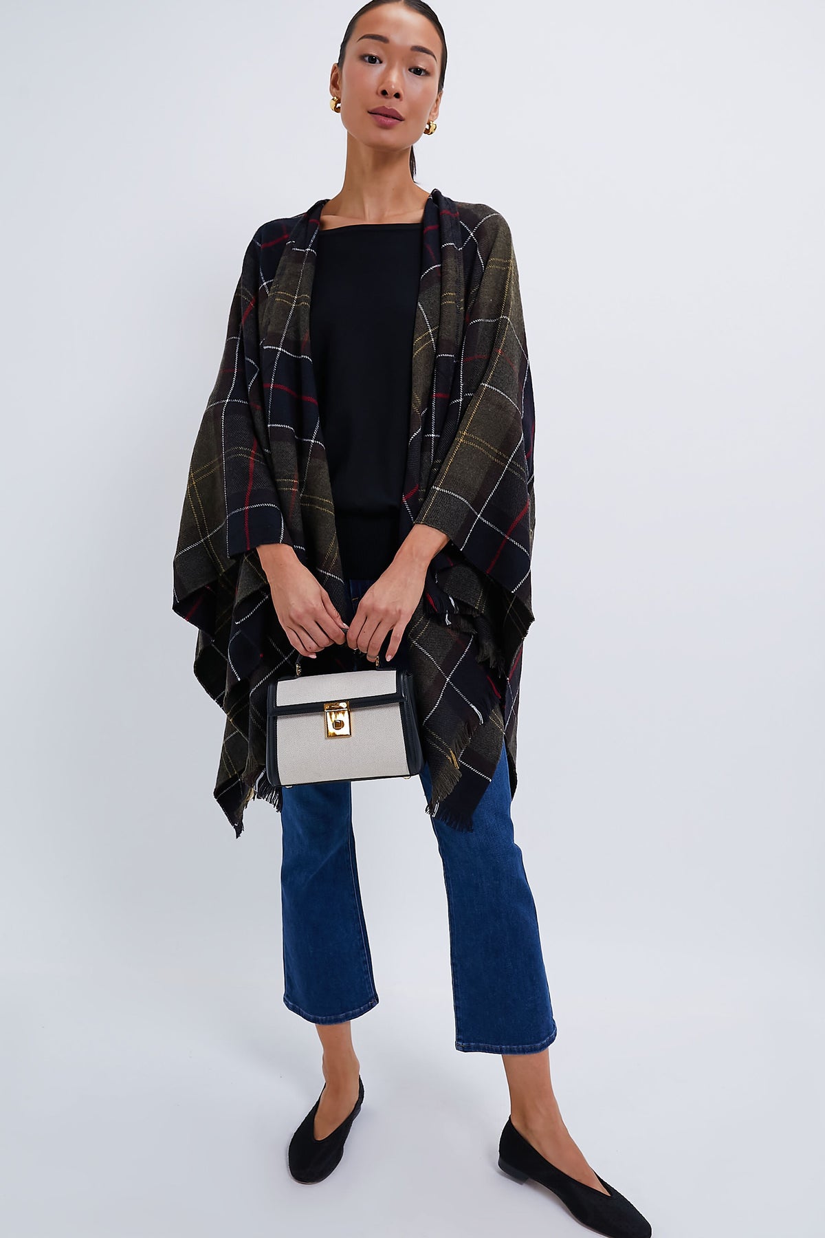 Woman wearing a Barbour tartan serape, black top, blue cropped jeans, black flats, and carrying a small handbag from Barbour.