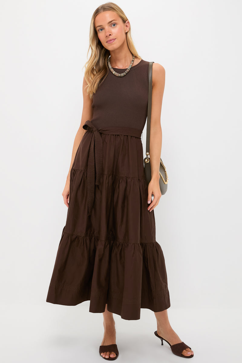 A woman models a sleeveless brown maxi A-line dress with a rib-knit bodice from Moon River, styled with a necklace, brown sandals, and a shoulder bag, standing against a plain background.