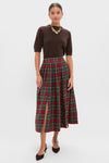 Woman in a brown short-sleeve top, Hunter Bell Bemelmans Plaid skirt with front slit, brown heels, and gold statement necklace stands against a plain light background—a perfect holiday party look.