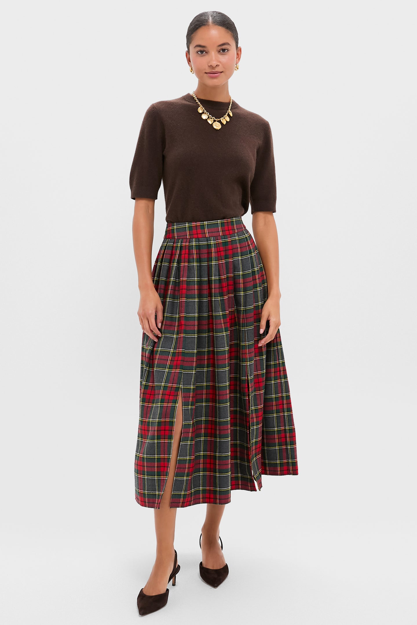 Woman in a brown short-sleeve top, Hunter Bell Bemelmans Plaid skirt with front slit, brown heels, and gold statement necklace stands against a plain light background—a perfect holiday party look.